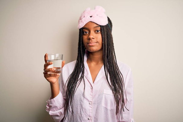 Молода афроамериканська жінка у піжамі та масці для очей випиває склянку води вночі з упевненим виразом розумного обличчя. - Фото, зображення