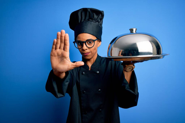 Νεαρή Αφροαμερικανή μαγείρισσα φορώντας στολή κρατώντας δίσκο σερβιτόρου με τρούλο με ανοιχτό χέρι κάνει στοπ με σοβαρή και σίγουρη έκφραση, χειρονομία άμυνας - Φωτογραφία, εικόνα
