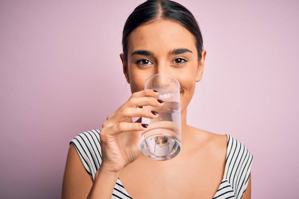 Νεαρή όμορφη μελαχρινή γυναίκα χαμογελά ευτυχισμένη και σίγουρη. Στέκεται με χαμόγελο στο πρόσωπο πίνοντας ένα ποτήρι υγιεινό νερό για να δροσιστεί σε απομονωμένο ροζ φόντο - Φωτογραφία, εικόνα