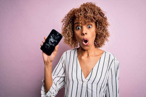 驚きの顔でショックで怖がって、恐怖の表情で恐怖と興奮を示す壊れたスマートフォンを持っている若いアフリカ系アメリカ人の巻き女性 - 写真・画像
