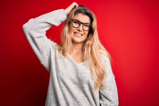 Young beautiful blonde woman wearing sweater and glasses over isolated red background confuse and wonder about question. Uncertain with doubt, thinking with hand on head. Pensive concept. - Fotografie, Obrázek