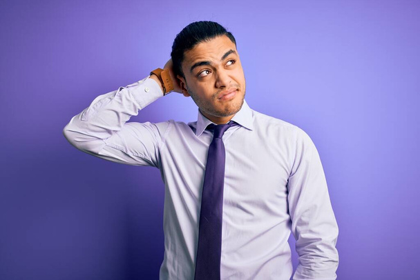 孤立した紫色の背景の上に立つエレガントなネクタイを身に着けている若いブラジルのビジネスマンは混乱し、質問について疑問に思う。疑いもなく、頭の上で手で考えて。包括的概念. - 写真・画像