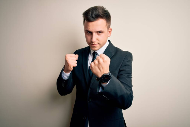 Молодой привлекательный бизнесмен в элегантном костюме и галстуке на изолированном фоне, готовый драться жестом кулачной защиты, сердитый и расстроенный, боится проблем
 - Фото, изображение