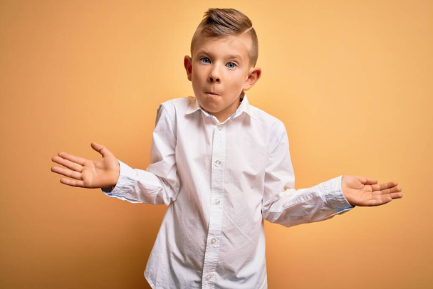 Young little caucasian kid with blue eyes wearing elegant white shirt over yellow background clueless and confused expression with arms and hands raised. Doubt concept. - Foto, Imagem