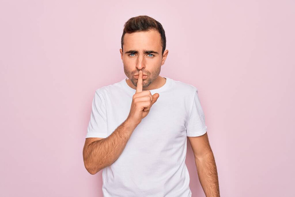 Handsome man with blue eyes wearing casual white t-shirt standing over pink background asking to be quiet with finger on lips. Silence and secret concept. - Photo, Image