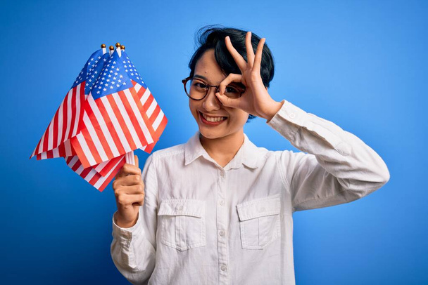 Junge schöne asiatische patriotische Mädchen mit Flaggen der Vereinigten Staaten feiern den Unabhängigkeitstag mit glücklichem Gesicht lächelnd tun ok Zeichen mit Hand auf Auge durch Finger schauen - Foto, Bild