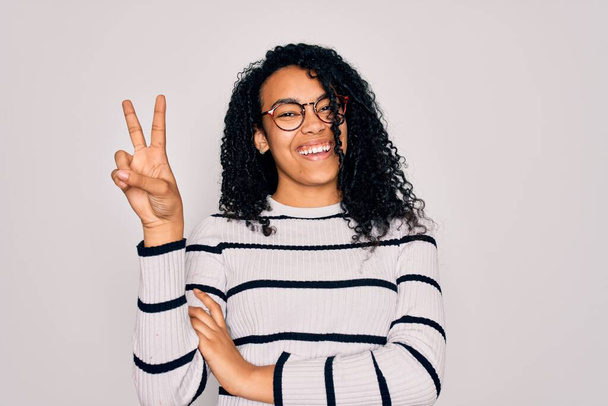 Mujer afroamericana joven con suéter a rayas y gafas sobre fondo blanco sonriendo con la cara feliz guiñando el ojo a la cámara haciendo señal de victoria. Número dos.
. - Foto, imagen