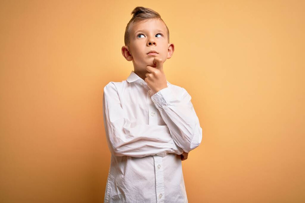 Young little caucasian kid with blue eyes wearing elegant white shirt over yellow background with hand on chin thinking about question, pensive expression. Smiling with thoughtful face. Doubt concept. - Photo, Image