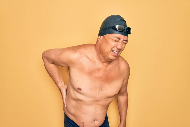 Hombre nadador de pelo gris de mediana edad con traje de baño, gorra y gafas Sufrimiento de dolor de espalda, tocando la espalda con la mano, dolor muscular
 - Foto, imagen