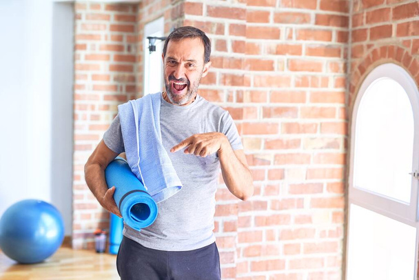 Среднего возраста красивый спортсмен держит мат и полотенце стоя перед делать упражнения в тренажерном зале очень счастливы указывая рукой и пальцем
 - Фото, изображение