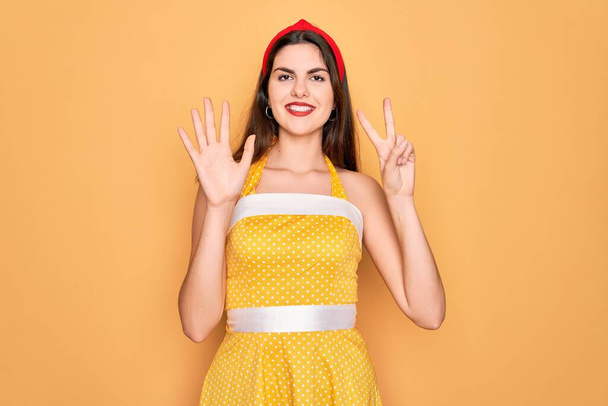 Joven hermosa pin up mujer con vestido vintage de moda de los años 50 sobre fondo amarillo que muestra y señala con los dedos número siete mientras sonríe confiado y feliz
. - Foto, Imagen