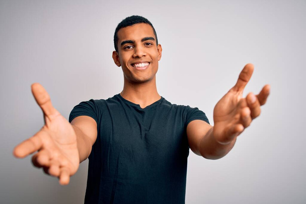 Jeune bel homme afro-américain portant un t-shirt décontracté debout sur fond blanc regardant la caméra souriant à bras ouverts pour un câlin. Expression joyeuse embrassant le bonheur
. - Photo, image