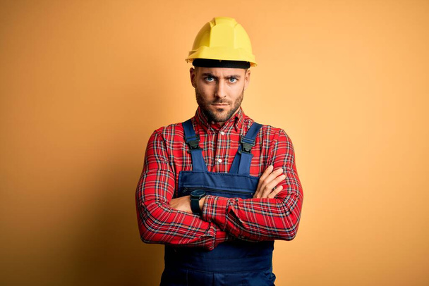 Молодой строитель в строительной форме и защитном шлеме на жёлтом изолированном фоне скептик и нервный, неодобрительное выражение лица со скрещенными руками. Отрицательное лицо
. - Фото, изображение