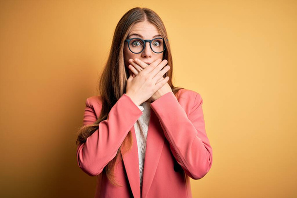 Young beautiful redhead woman wearing jacket and glasses over isolated yellow background shocked covering mouth with hands for mistake. Secret concept. - Foto, Bild