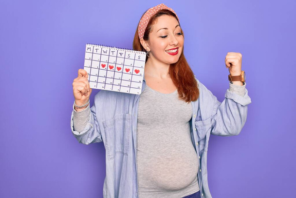 Молодая красивая рыжая беременная женщина ожидает ребенка период проведения календаря указывая и показывая с большим пальцем в сторону со счастливым лицом улыбаясь
 - Фото, изображение