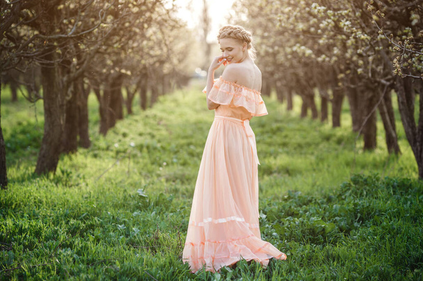 κορίτσι με ξανθά μαλλιά σε ένα ελαφρύ φόρεμα στον ανθισμένο κήπο. έννοια της γυναικείας ανοιξιάτικης μόδας. Νεαρό μοντέλο κοιτάζοντας φωτογραφική μηχανή εκφραστική. - Φωτογραφία, εικόνα