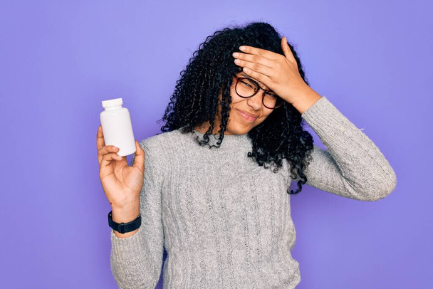 Young african american curly woman holding pills standing over isolated purple background stressed with hand on head, shocked with shame and surprise face, angry and frustrated. Fear and upset for mistake. - 写真・画像