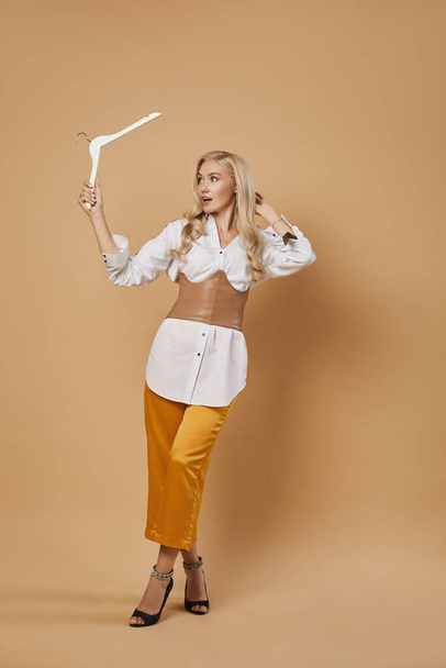Beautiful young woman in fashionable outfit holding an empty wooden hanger in her hand and expressing that nothing to wear. Shopping concept. Beauty concept. Isolated with copy space. - Photo, image