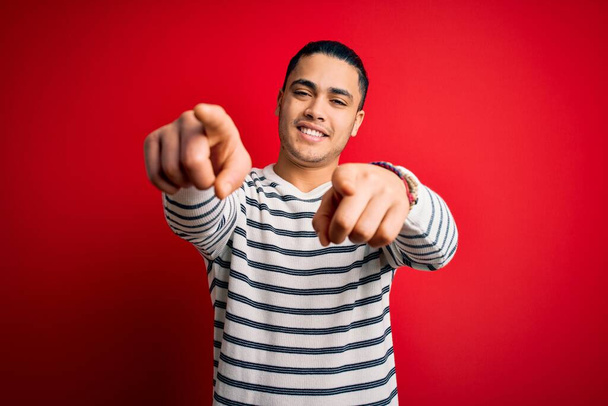 Junger brasilianischer Mann in lässigem gestreiftem T-Shirt steht über isoliertem rotem Hintergrund und zeigt mit den Fingern auf dich und die Kamera, lächelt positiv und fröhlich - Foto, Bild