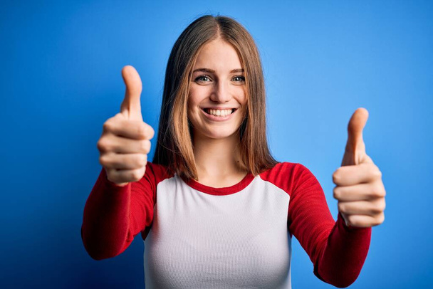Young beautiful redhead woman wearing casual t-shirt over isolated blue background approving doing positive gesture with hand, thumbs up smiling and happy for success. Winner gesture. - Photo, Image