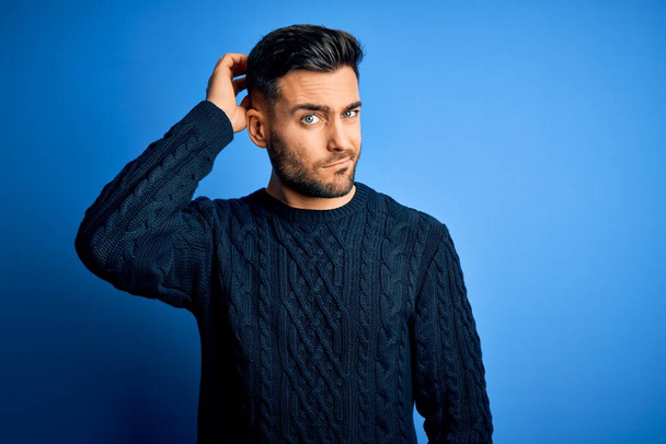 Young handsome man wearing casual sweater standing over isolated blue background confuse and wondering about question. Uncertain with doubt, thinking with hand on head. Pensive concept. - Photo, Image