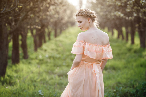 dziewczyna z blond włosami w jasnej sukience w kwitnącym ogrodzie. koncepcja kobiecej wiosennej mody. Młody model patrzący na aparat ekspresyjny. - Zdjęcie, obraz