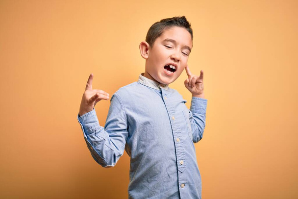 Niño pequeño con camisa elegante de pie sobre fondo aislado amarillo gritando con expresión loca haciendo símbolo de roca con las manos arriba. Estrella de la música. Concepto pesado
. - Foto, Imagen