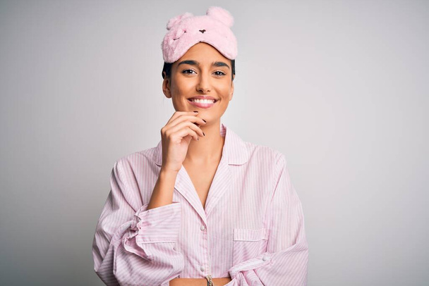 Young beautiful brunette woman wearing pajama and sleep mask over white background looking confident at the camera with smile with crossed arms and hand raised on chin. Thinking positive. - Photo, Image