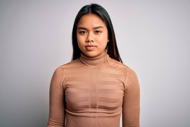 Young beautiful asian girl wearing casual turtleneck sweater standing over white background with serious expression on face. Simple and natural looking at the camera. - Photo, Image