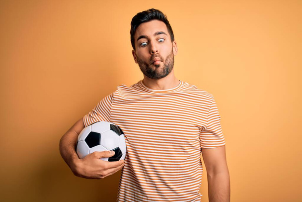 Handsome player man with beard playing soccer holding footballl ball over yellow background making fish face with lips, crazy and comical gesture. Funny expression. - Photo, Image