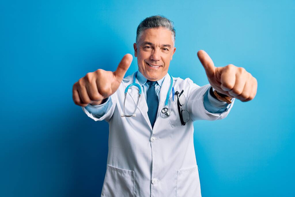 Medioevo bell'uomo medico dai capelli grigi che indossa cappotto e stetoscopio blu che approva facendo gesto positivo con la mano, pollice in su sorridente e felice per il successo. Gesto del vincitore
. - Foto, immagini
