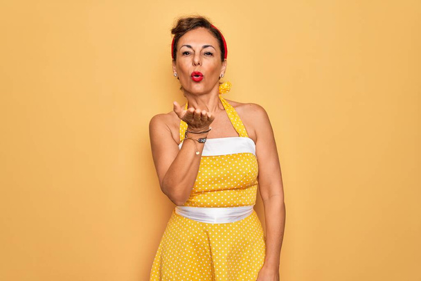 Μέση ηλικία ανώτερος pin up γυναίκα φορώντας ρετρό στυλ 50s φόρεμα πάνω από κίτρινο φόντο κοιτάζοντας την κάμερα φυσάει ένα φιλί με το χέρι στον αέρα είναι όμορφη και σέξι. Έκφραση αγάπης. - Φωτογραφία, εικόνα