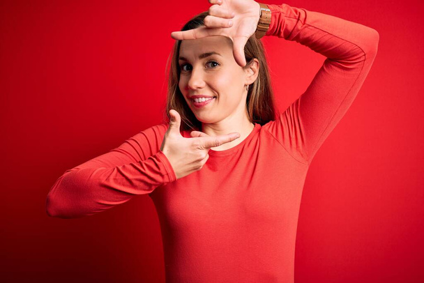 Jonge mooie blonde vrouw draagt casual t-shirt staan over geïsoleerde rode achtergrond glimlachen maken frame met handen en vingers met gelukkig gezicht. Creativiteit en fotografie concept. - Foto, afbeelding