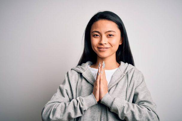 Jovem mulher esportiva chinesa bonita vestindo camisola sobre fundo branco isolado orando com as mãos juntas pedindo perdão sorrindo confiante
. - Foto, Imagem