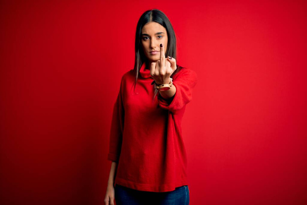 Молодая красивая брюнетка в обычном свитере на изолированном красном фоне показывает средний палец, невежливый и грубый отъебись выражение лица
 - Фото, изображение