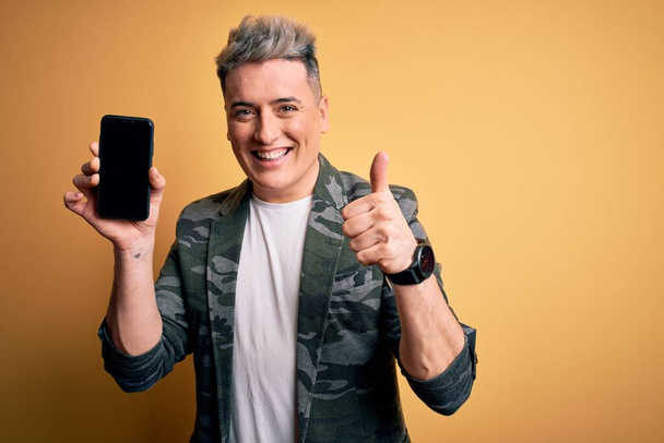 Νεαρός σύγχρονος άνθρωπος δείχνει το smartphone κενή οθόνη πάνω από το κίτρινο απομονωμένο φόντο χαρούμενος με μεγάλο χαμόγελο κάνει ok σημάδι, τον αντίχειρα επάνω με τα δάχτυλα, εξαιρετικό σημάδι - Φωτογραφία, εικόνα
