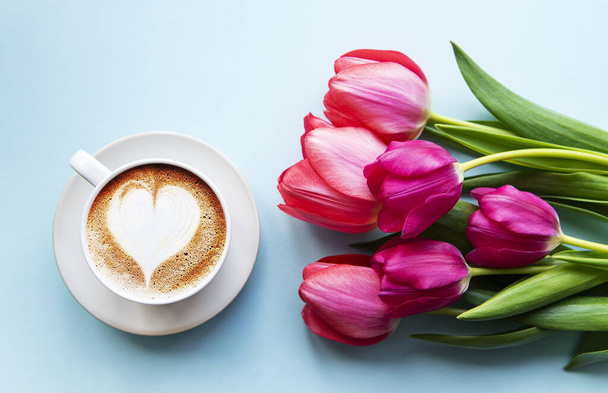 Coffee cup with latte art and pink tulips on the blue background. Top view, flat lay.  - Photo, Image