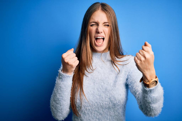 Young beautiful redhead woman wearing casual sweater over isolated blue background angry and mad raising fists frustrated and furious while shouting with anger. Rage and aggressive concept. - Foto, afbeelding