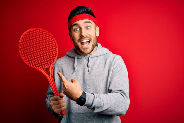 Tenis raketi tutan yakışıklı genç sporcu kırmızı arka planda spor giysisi giyiyor. Elle ve parmakla göstererek çok mutlu. - Fotoğraf, Görsel