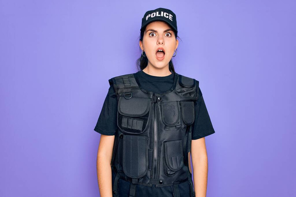Fiatal rendőr nő visel biztonsági golyóálló mellény egyenruha felett lila háttér félek, és sokkolt a meglepetés és csodálkozó kifejezés, félelem és izgatott arc. - Fotó, kép