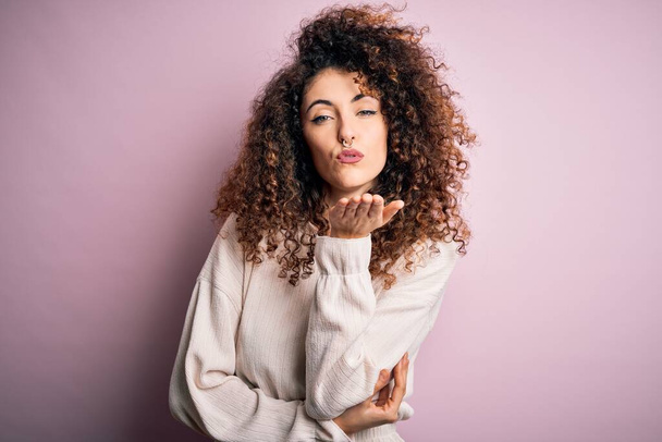 Beautiful woman with curly hair and piercing wearing casual sweater over pink background looking at the camera blowing a kiss with hand on air being lovely and sexy. Love expression. - Photo, Image