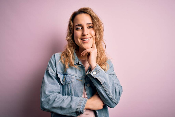 Young beautiful blonde woman wearing casual denim jacket standing over pink background looking confident at the camera with smile with crossed arms and hand raised on chin. Thinking positive. - Φωτογραφία, εικόνα