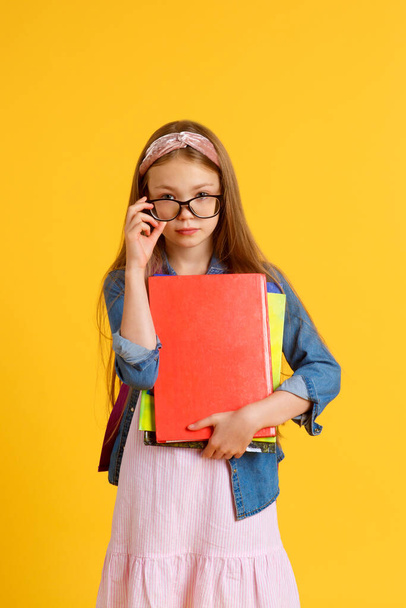 portrait of happy caucasian school girl with a book. beautiful positive girl with long red hair stand and hug her book, look at camera. she is preparing to school . isolated yellow background - Fotó, kép