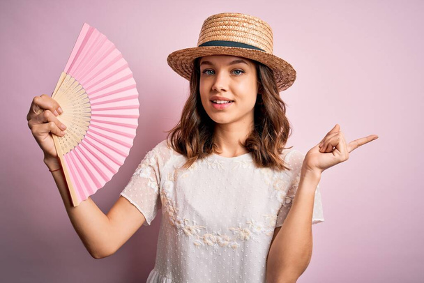 Jeune fille de mode blonde utilisant un ventilateur de main espagnol en bois sur une chaude journée d'été très heureux pointant avec la main et le doigt sur le côté
 - Photo, image