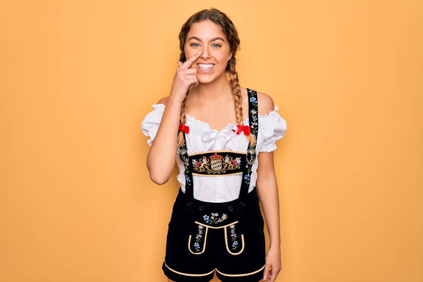 青い目をした若い美しいブロンドのドイツ人女性は、伝統的な10月祭のドレスを着て顔と鼻に手の指で指して、陽気に笑っています。美の概念 - 写真・画像