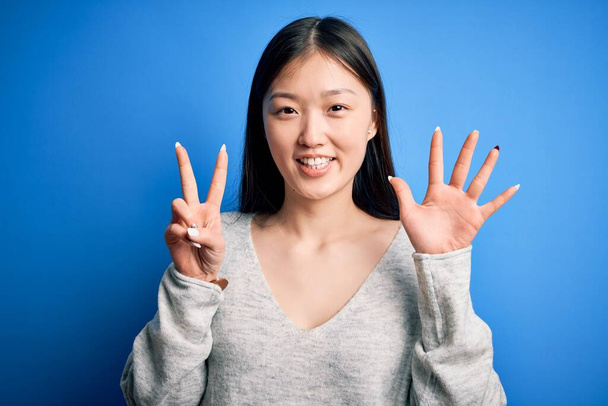 Fiatal gyönyörű ázsiai nő, alkalmi pulóvert visel, kék elszigetelt háttér felett áll, mutatva és mutatva fel a hetes számú ujjaival, miközben magabiztosan és boldogan mosolyog.. - Fotó, kép