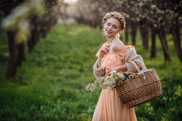 girl with blonde hair in a light dress in flowering garden. girl in a beautiful dress and knitted sweater enjoys the sunset in a pear flowering garden, with a basket of flowers - Fotoğraf, Görsel