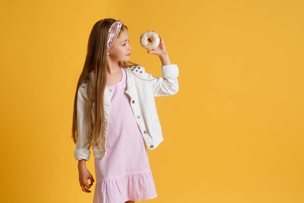 Crazy cheerful blonde girl in birthday hat smiling, having fun and looking through two red donuts on her eyes. Sweets. Yellow studio background. - Foto, imagen
