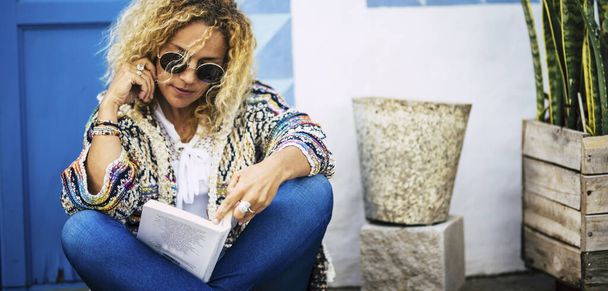 Красива блондинка модель доросла жінка насолоджується книгою читання на відкритому повітрі з синіми дверима і білим будинком на задньому плані
  - Фото, зображення
