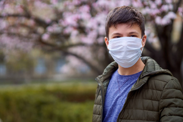 jongen met gezichtsmasker is in de stad buiten, bloeiende bomen, voorjaarsseizoen, bloei tijd - concept van allergieën en gezondheid bescherming tegen stoffige lucht - Foto, afbeelding
